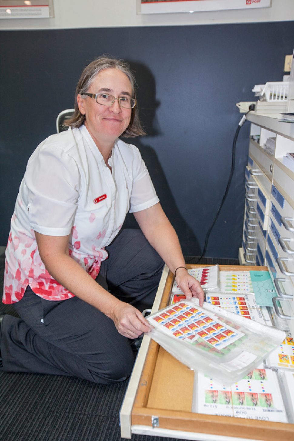 Melissa Anderson kneeling down behind a desk, looking at a tray of stamps